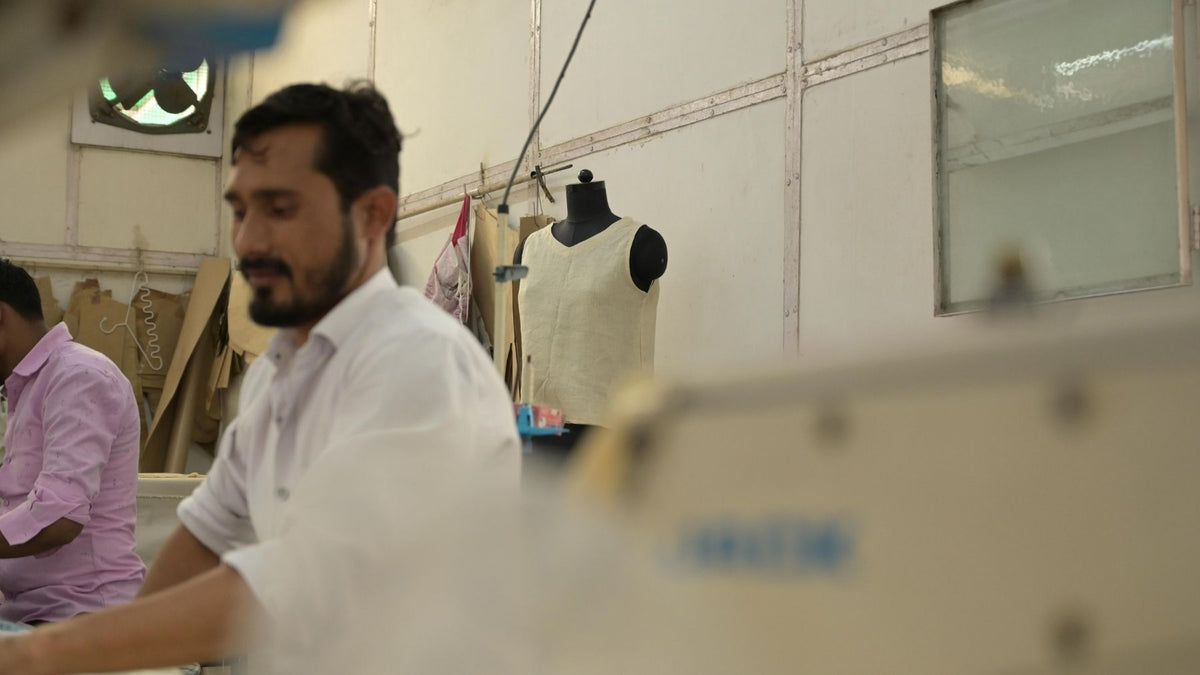 A man in a white shirt works at a sewing machine in a workshop, with a mannequin wearing a pearl-colored garment and other sewing equipment visible in the background.