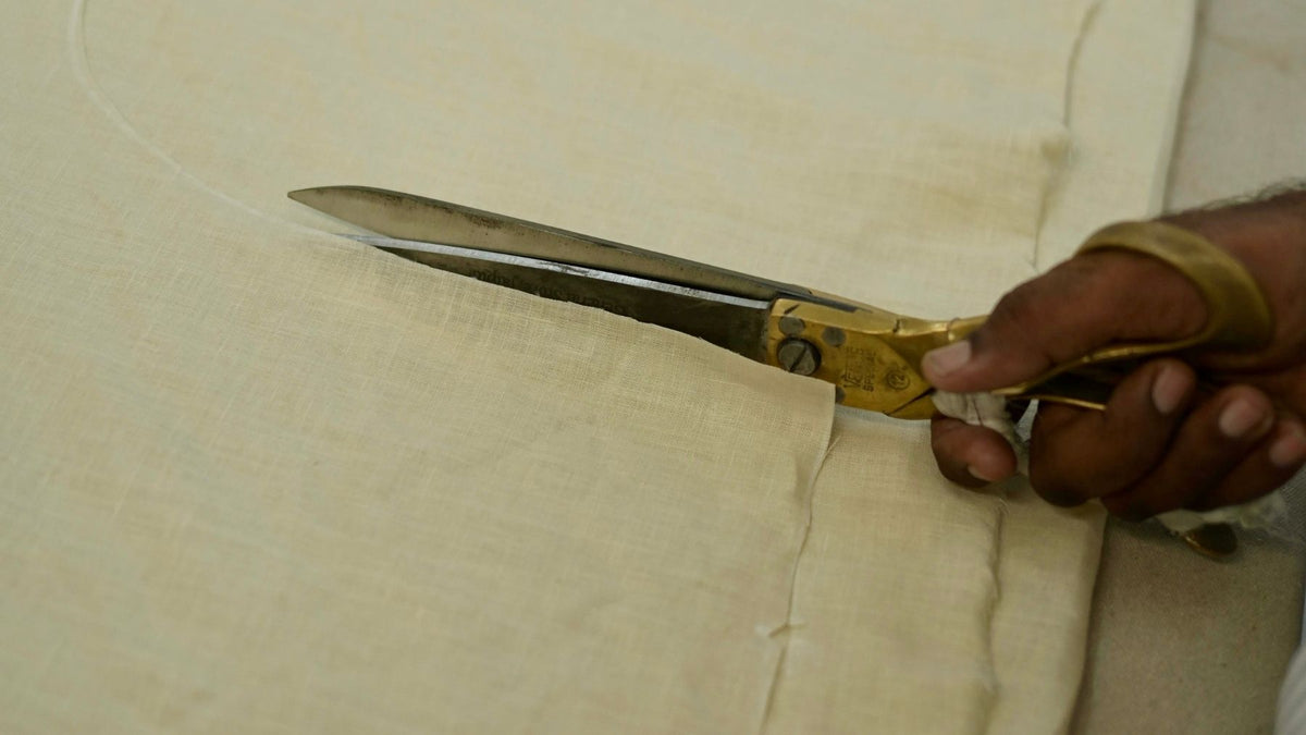 A close-up of a hand holding a pair of brass and steel scissors while cutting a pearl linen fabric.