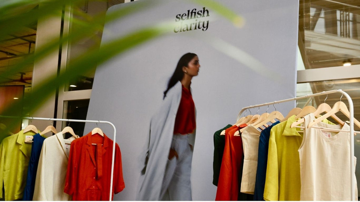 A clothing display in a retail store featuring a large vertical sign with the brand name 'selfish clarity' and a model photo, alongside hanging garments in clean expressive colors on both sides.