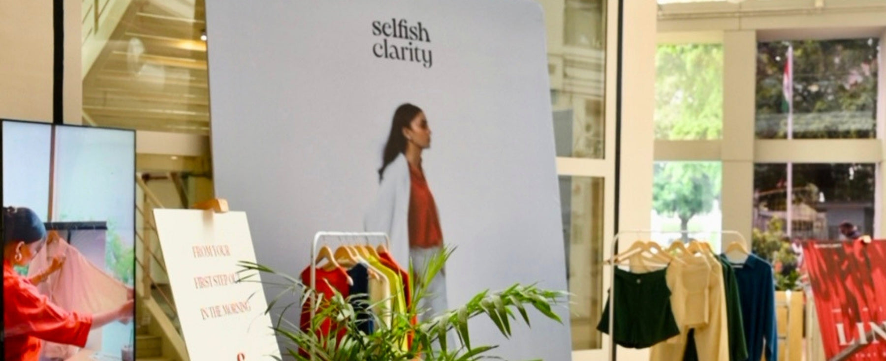 A retail store interior with a large vertical sign reading 'selfish clarity' behind a model photo, clothing racks with garments, a plant in the foreground, and window views of the street outside.
